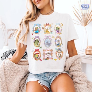 Puede incluir: Camiseta blanca con personajes de dibujos animados enmarcados. Los marcos son de varios colores y dise&ntilde;os, con personajes como un oso, una princesa y un cocodrilo. La camiseta tiene el texto "COMIC DESIGNS" en la esquina superior derecha.