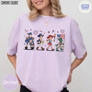 May include: Orchid-colored t-shirt with a patriotic cartoon design. The graphic features four cartoon characters marching with fireworks and flags. The shirt has the words "COMFORT COLORS" and "COMIC DESIGNS" printed on it.