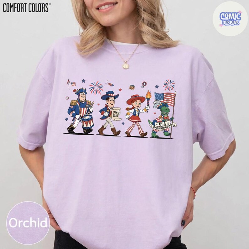 May include: Orchid-colored t-shirt with a patriotic cartoon design. The graphic features four cartoon characters marching with fireworks and flags. The shirt has the words "COMFORT COLORS" and "COMIC DESIGNS" printed on it.