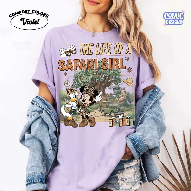 May include: Violet Comfort Colors t-shirt with a safari theme. The shirt features the text "THE LIFE OF A SAFARI GIRL" and a graphic of Minnie and Daisy Duck in safari outfits. The shirt also has a graphic of a safari vehicle and a tree.