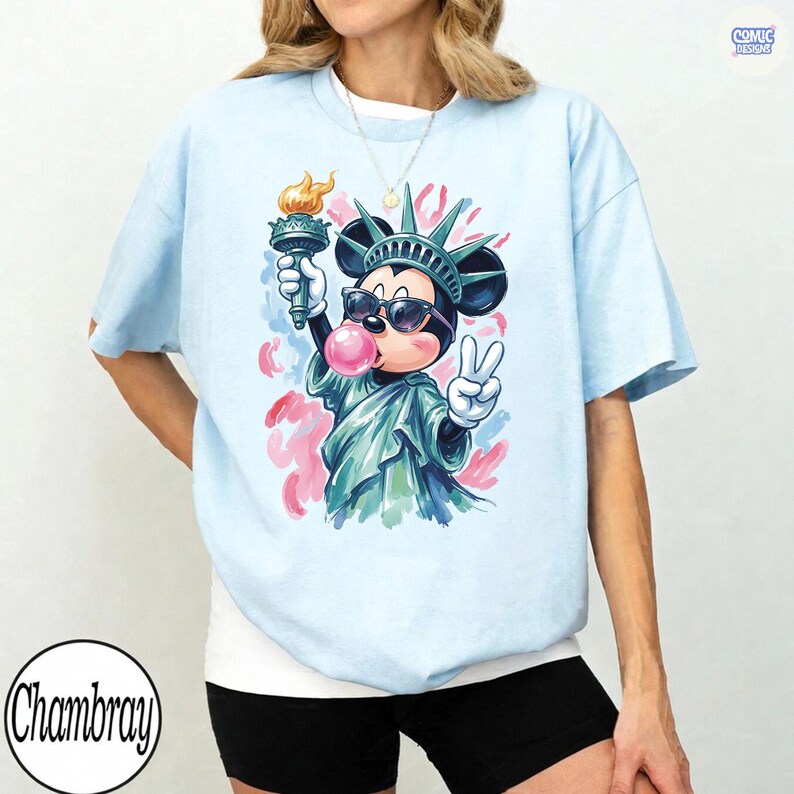 May include: A light blue t-shirt featuring a graphic of a cartoon mouse dressed as the Statue of Liberty, wearing sunglasses and blowing a pink bubble. The mouse holds a torch and makes a peace sign, set against a background of abstract pink and red brushstrokes.