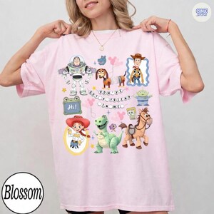 May include: A light pink t-shirt featuring a graphic print of various Toy Story characters including Buzz Lightyear, Woody, Jessie, Rex, Slinky Dog, and Bullseye, with the text "YOU'VE GOT A FRIEND IN ME" and a small green sign that says "Hi!"