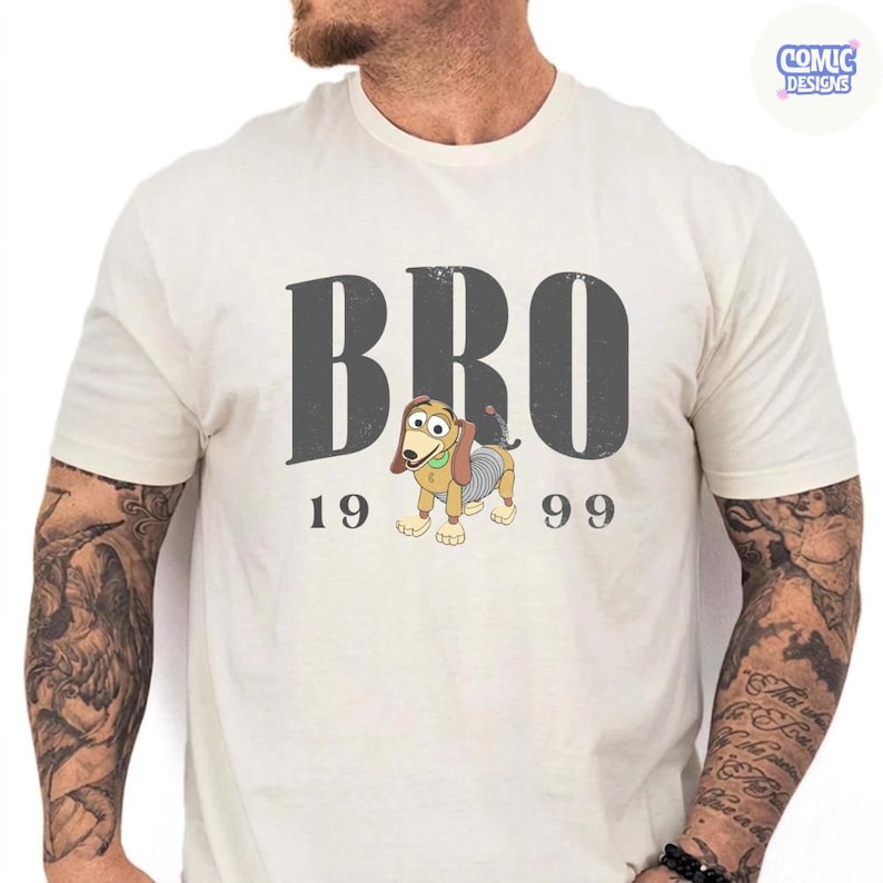 K&ouml;nnte beinhalten: Cremefarbenes T-Shirt mit dem Wort "BRO" in Dunkelgrau und den Zahlen "19" und "99" darunter. In der Mitte befindet sich ein Cartoon-Hund mit einem federartigen K&ouml;rper. Das Shirt hat kurze &Auml;rmel und einen Rundhalsausschnitt.