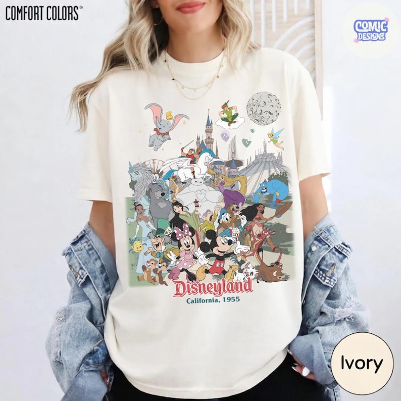 Puede incluir: Camiseta de color marfil con un gr&aacute;fico colorido de Disneyland. El dise&ntilde;o incluye varios personajes de Disney, un castillo y las palabras "Disneyland California, 1955". La camiseta es de Comfort Colors y tiene un logotipo de Comic Designs.