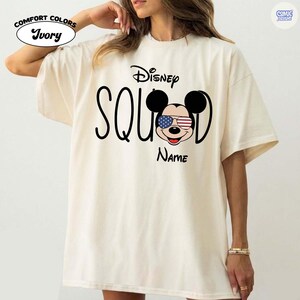May include: Ivory-colored t-shirt with the text "Disney Squad" and a Mickey Mouse graphic wearing American flag sunglasses. The shirt also has the word "Name" below the graphic. The shirt has the "Comfort Colors" logo.