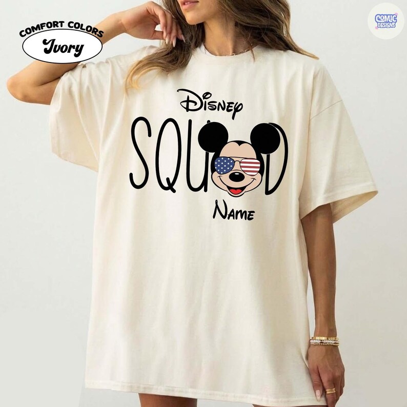 May include: Ivory-colored t-shirt with the text "Disney Squad" and a Mickey Mouse graphic wearing American flag sunglasses. The shirt also has the word "Name" below the graphic. The shirt has the "Comfort Colors" logo.