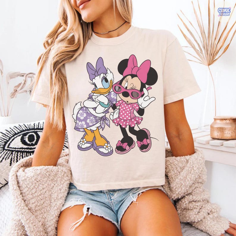 Puede incluir: Una camiseta de manga corta color crema con un estampado gr&aacute;fico de Daisy Duck y Minnie Mouse. Daisy lleva un pareo y un lazo con estampado floral morado, mientras que Minnie viste un vestido rosa de lunares, un lazo rosa y gafas de sol rosas.