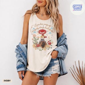 Op de afbeelding: Een ivoorkleurige mouwloze tanktop met een grafische print van een cowgirl op een paard, omringd door cactussen en bloemen. De tekst luidt, Love has ways of bringing things back to life. Een lichtblauw spijkerhemd hangt over de schouders.
