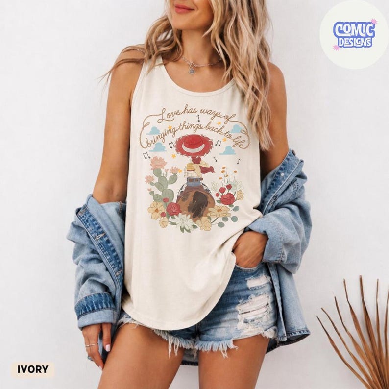 Op de afbeelding: Een ivoorkleurige mouwloze tanktop met een grafische print van een cowgirl op een paard, omringd door cactussen en bloemen. De tekst luidt, Love has ways of bringing things back to life. Een lichtblauw spijkerhemd hangt over de schouders.
