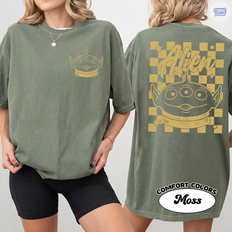 Puede incluir: Camiseta verde oliva con un gr&aacute;fico de alien&iacute;gena dorado en la parte delantera y trasera. La parte trasera presenta la palabra "Alien" encima del gr&aacute;fico del alien&iacute;gena, sobre un fondo a cuadros dorados y beige. La camiseta est&aacute; etiquetada como "Comfort Colors Moss".