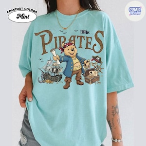 May include: Mint-colored t-shirt with a pirate-themed graphic. The design features a cartoon bear dressed as a pirate, a pirate ship, treasure chest, and the word "Pirates" in brown letters. The shirt has the words "Comfort Colors" and "Mint" printed on it.