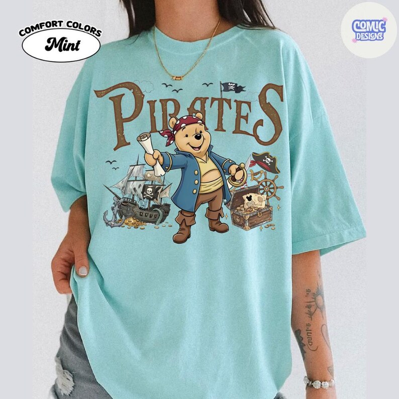 May include: Mint-colored t-shirt with a pirate-themed graphic. The design features a cartoon bear dressed as a pirate, a pirate ship, treasure chest, and the word "Pirates" in brown letters. The shirt has the words "Comfort Colors" and "Mint" printed on it.