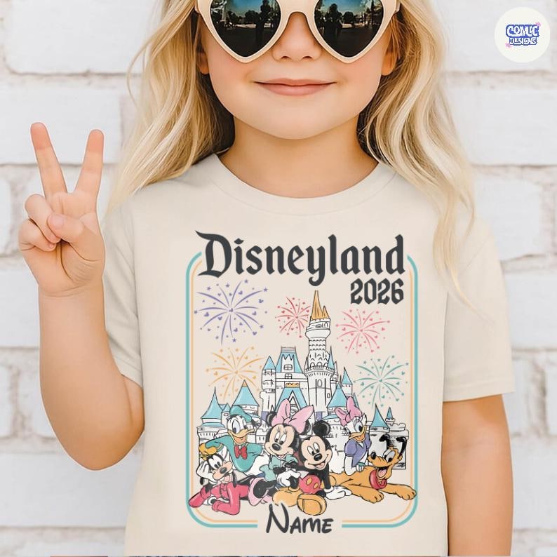 Op de afbeelding: Een beige t-shirt met de tekst "Disneyland 2026" en een kleurrijke afbeelding van een kasteel, vuurwerk en Disney-figuren. Het shirt heeft onderaan een gepersonaliseerde "Name"-sectie.