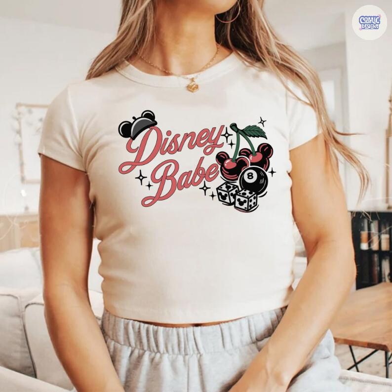 May include: Cream-coloured crop top featuring the text "Disney Babe" in pink script, with black star accents. The graphic includes Mickey Mouse ears, cherries, dice, and an 8-ball.