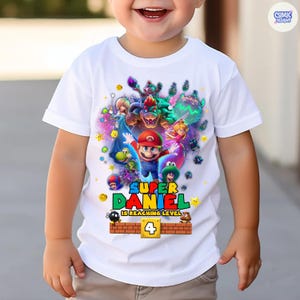 Pode incluir: Camiseta branca com um design colorido de personagens do Super Mario. O design inclui o texto "SUPER DANIEL IS REACHING LEVEL 4". A camiseta &eacute; para uma crian&ccedil;a.