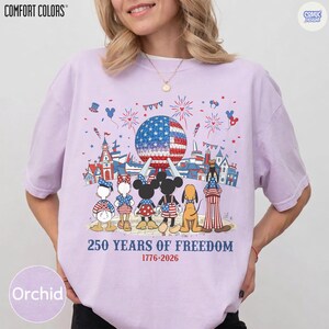 May include: A light purple t-shirt featuring a graphic of cartoon characters standing before a large geodesic dome and castle. Above them are fireworks and the text 250 YEARS OF FREEDOM 1776-2026. The characters wear patriotic red, white, and blue clothing.