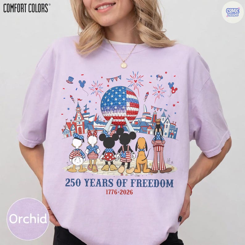 May include: A light purple t-shirt featuring a graphic of cartoon characters standing before a large geodesic dome and castle. Above them are fireworks and the text 250 YEARS OF FREEDOM 1776-2026. The characters wear patriotic red, white, and blue clothing.