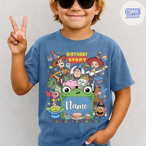 May include: A blue short-sleeve t-shirt featuring a colorful graphic of various animated toy characters wearing party hats. The design includes the text Birthday Story and a central green frame with the word Name above a birthday cake illustration.