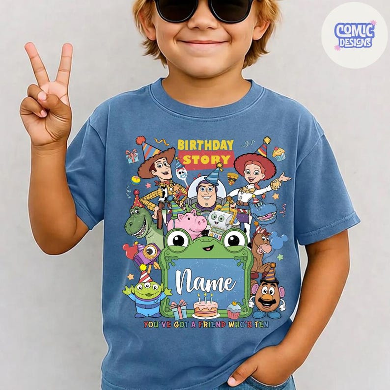 May include: A blue short-sleeve t-shirt featuring a colorful graphic of various animated toy characters wearing party hats. The design includes the text Birthday Story and a central green frame with the word Name above a birthday cake illustration.