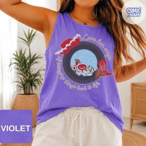 May include: A violet tank top featuring a graphic of a black tire framing a blue sky scene with a cowboy hat, a red cowboy boot, and small character figures. The text reads, Love has ways of bringing things back to life, in a cursive font.