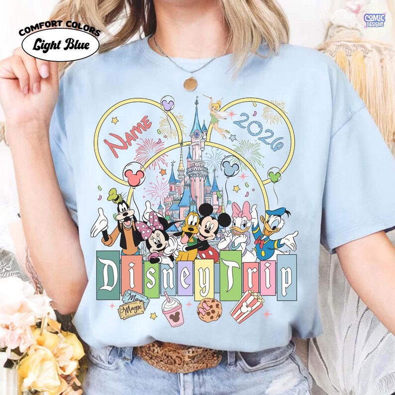May include: Light blue t-shirt with a Disney Trip design. The design features Disney characters, a castle, fireworks, and the text "Name" and "2026". The shirt also has the words "Comfort Colors" and "Light Blue" printed on it.