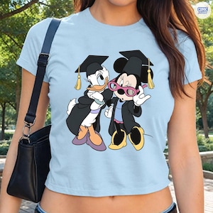 Puede incluir: Camiseta azul claro con un dise&ntilde;o de Daisy y Minnie Mouse vestidas con atuendos de graduaci&oacute;n. Los personajes de dibujos animados llevan birretes y togas negras con borlas doradas. La camiseta tiene cuello redondo y mangas cortas.