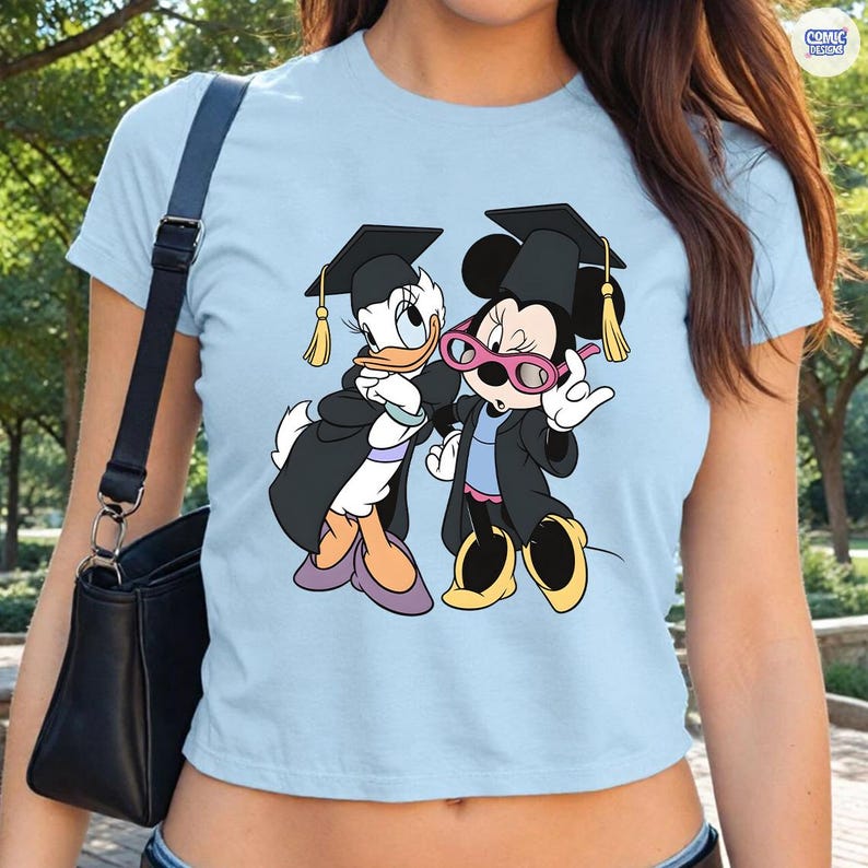 Puede incluir: Camiseta azul claro con un dise&ntilde;o de Daisy y Minnie Mouse vestidas con atuendos de graduaci&oacute;n. Los personajes de dibujos animados llevan birretes y togas negras con borlas doradas. La camiseta tiene cuello redondo y mangas cortas.