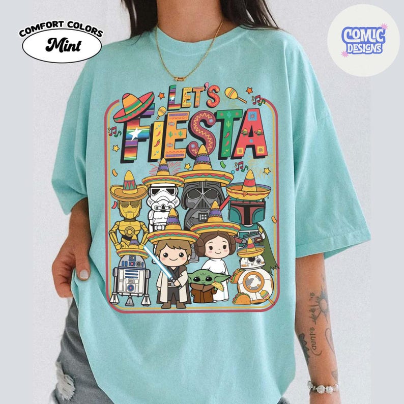 May include: Mint-colored t-shirt with a graphic that says "Let's Fiesta." The graphic features cartoon characters wearing sombreros. The shirt has the words "Comfort Colors" and "Comic Designs" printed on it.