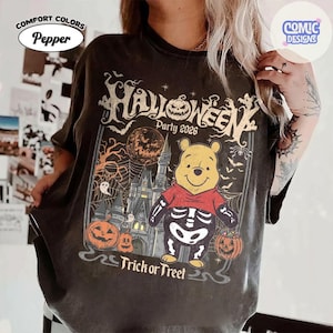 Puede incluir: Una camiseta color pimienta con un gr&aacute;fico de un oso amarillo con disfraz de esqueleto que sostiene un cubo de calabaza. El dise&ntilde;o incluye un castillo, una luna llena, telara&ntilde;as, murci&eacute;lagos, fantasmas y el texto "Halloween Party 2026 Trick or Treet".