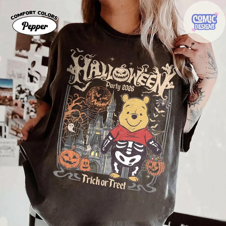 Puede incluir: Una camiseta color pimienta con un gr&aacute;fico de un oso amarillo con disfraz de esqueleto que sostiene un cubo de calabaza. El dise&ntilde;o incluye un castillo, una luna llena, telara&ntilde;as, murci&eacute;lagos, fantasmas y el texto "Halloween Party 2026 Trick or Treet".