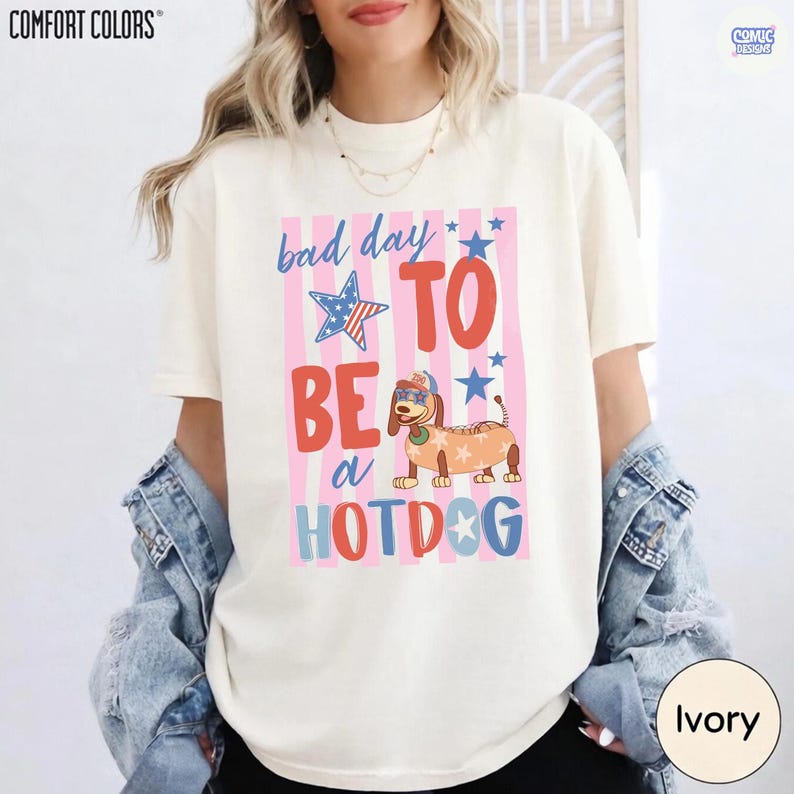 May include: Ivory-colored Comfort Colors t-shirt with a graphic that reads "Bad Day To Be a Hotdog." The design features a dachshund wearing sunglasses and a hotdog costume, set against a pink and white striped background with stars.