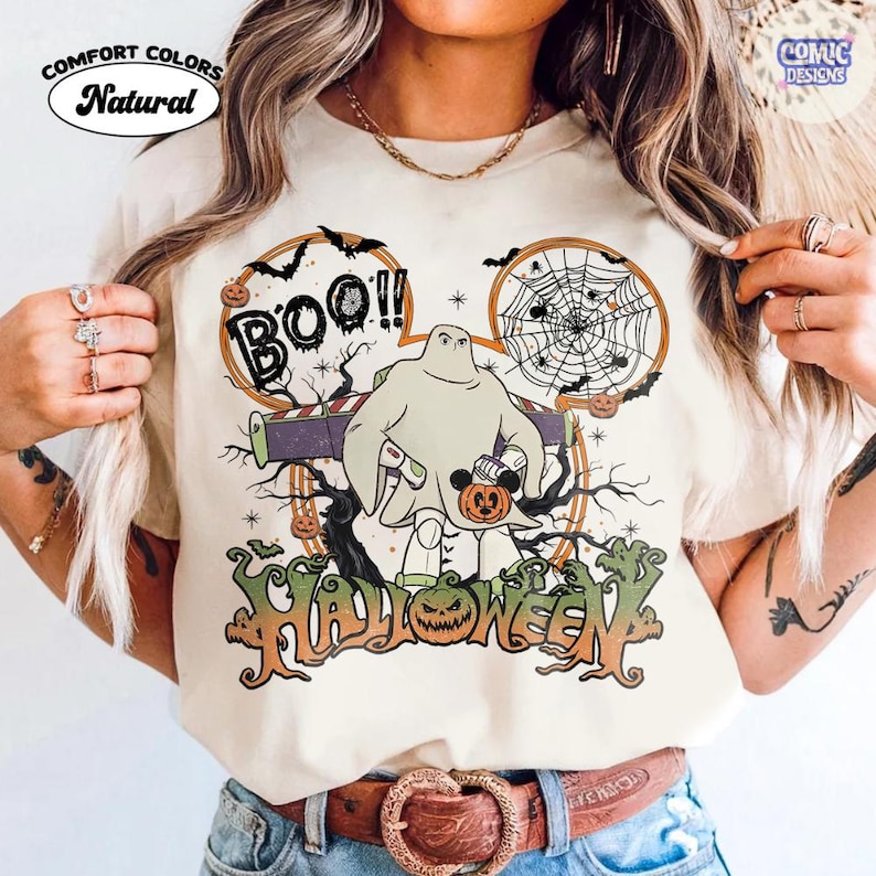 Peut inclure: Un t-shirt de couleur naturelle avec un motif repr&eacute;sentant un personnage d&eacute;guis&eacute; en fant&ocirc;me portant une combinaison de ranger de l'espace. Le design inclut le texte "BOO!!" et "HALLOWEEN", une toile d'araign&eacute;e, des chauves-souris et une citrouille.