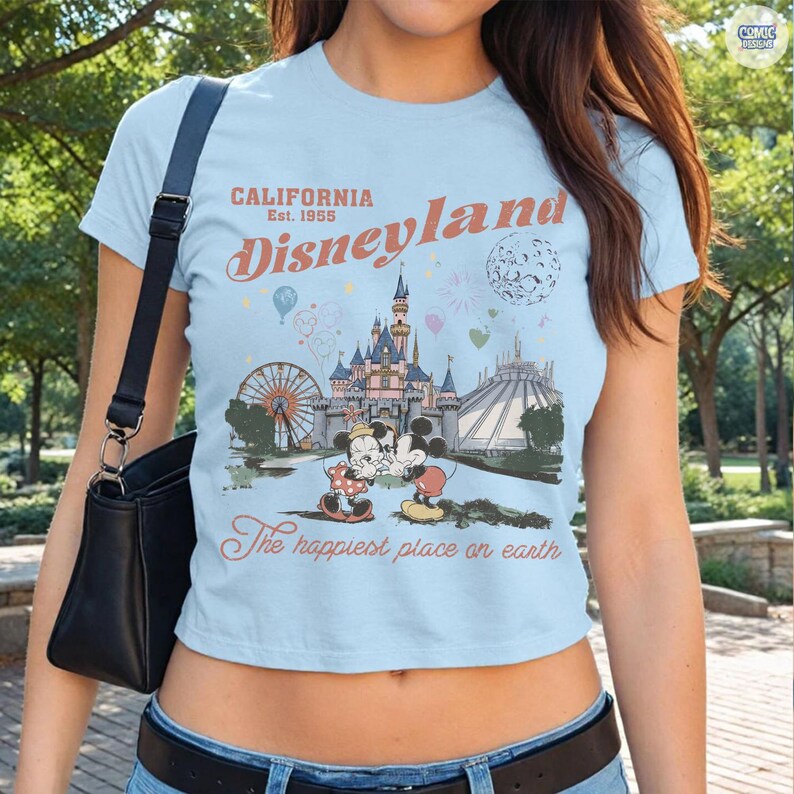 Op de afbeelding: Lichtblauw crop top met een Disneyland-afbeelding. Het ontwerp bevat de tekst "California Disneyland Est. 1955" en een illustratie van het kasteel, Mickey en Minnie Mouse en andere parkattracties. De zin "The happiest place on earth" is ook opgenomen.