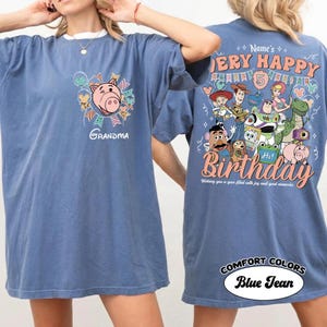 May include: Blue jean Comfort Colors t-shirt with a graphic of a pig and the word "Grandma" on the front. The back features a "Very Happy Birthday" design with cartoon characters and the text "Wishing you a year filled with joy and good memories."