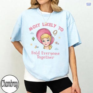 May include: A light blue short-sleeve t-shirt featuring a graphic of a blonde character in a pink bonnet. The text reads Most Likely To Hold Everyone Together, surrounded by small icons of a cactus, stars, a rocket, and building blocks.