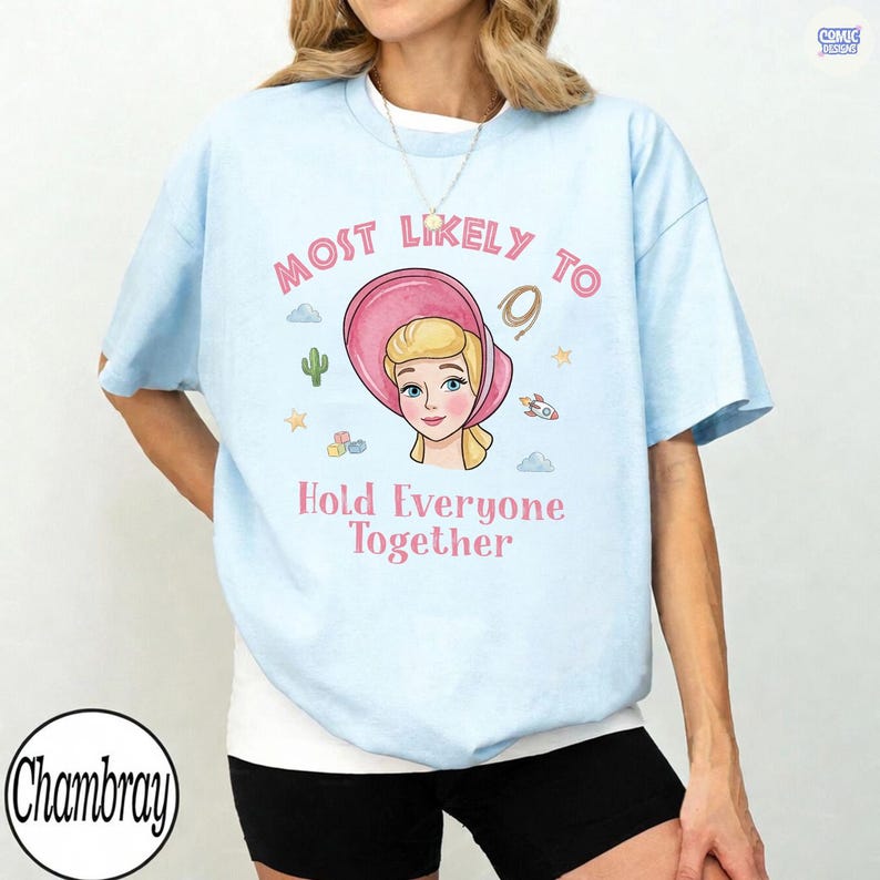 May include: A light blue short-sleeve t-shirt featuring a graphic of a blonde character in a pink bonnet. The text reads Most Likely To Hold Everyone Together, surrounded by small icons of a cactus, stars, a rocket, and building blocks.