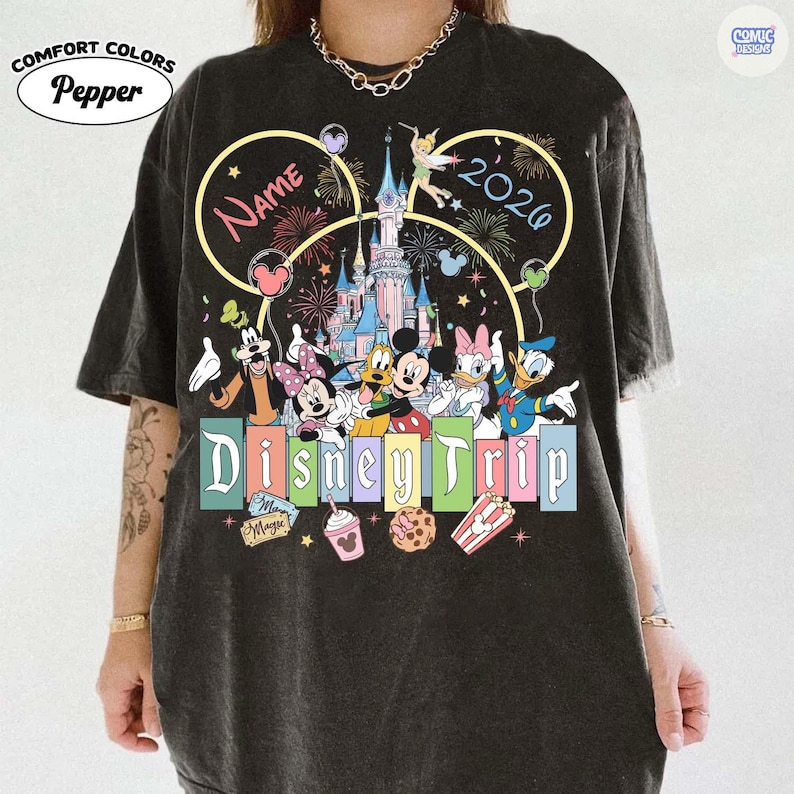 May include: Dark gray Comfort Colors t-shirt featuring a colorful Disney Trip design. The design includes Mickey and Minnie Mouse, Donald and Daisy Duck, and Goofy. The text "Name" and "2026" are also included.