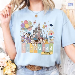 May include: Light blue t-shirt featuring a colorful graphic of Disney characters, a castle, balloons, and the text "Congratulations". Below, the words "PreK Grad Mommy" are displayed. The shirt has a crew neck and short sleeves.