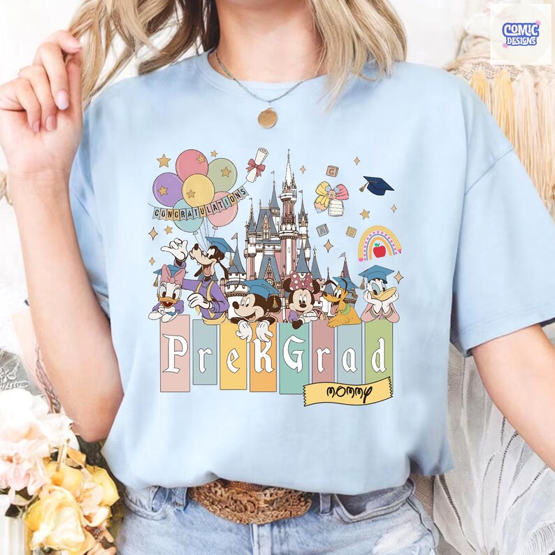 May include: Light blue t-shirt featuring a colorful graphic of Disney characters, a castle, balloons, and the text "Congratulations". Below, the words "PreK Grad Mommy" are displayed. The shirt has a crew neck and short sleeves.