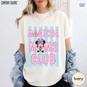 K&ouml;nnte beinhalten: Elfenbeinfarbenes T-Shirt mit dem Text "MAGICAL MOMS CLUB" in Pink, mit einer Minnie Mouse-Grafik und einer Schloss-Illustration. Das Design befindet sich auf einem hellblau gestreiften Hintergrund. Marke Comfort Colors.