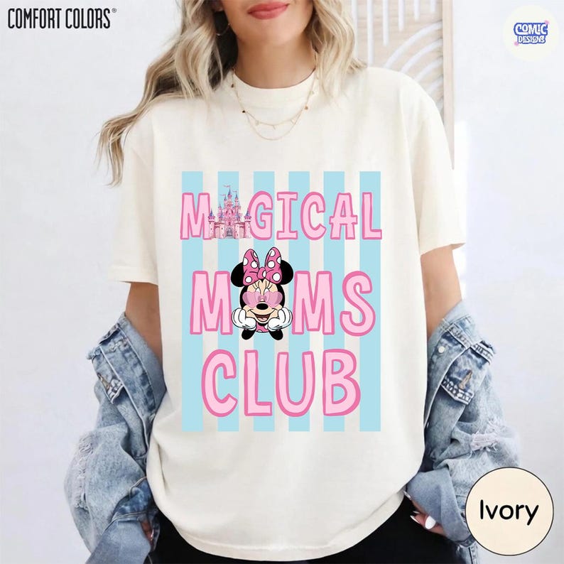 K&ouml;nnte beinhalten: Elfenbeinfarbenes T-Shirt mit dem Text "MAGICAL MOMS CLUB" in Pink, mit einer Minnie Mouse-Grafik und einer Schloss-Illustration. Das Design befindet sich auf einem hellblau gestreiften Hintergrund. Marke Comfort Colors.
