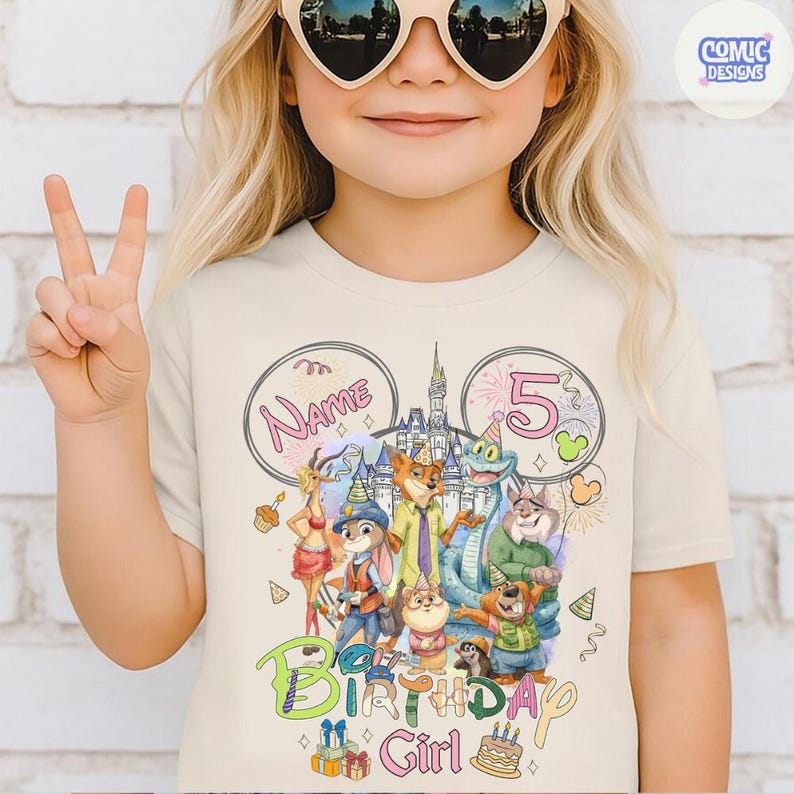 以下が含まれることがあります： クリーム色のTシャツ。カラフルなグラフィックデザインが特徴です。「Birthday Girl」の文字と数字の「5」、様々な漫画のキャラクター、お城が描かれています。「Name」の文字も含まれています。
