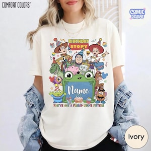 May include: An ivory t-shirt featuring a colorful graphic print of various animated characters wearing party hats. The design includes the text "BIRTHDAY STORY", "Name", and "YOU'VE GOT A FRIEND WHO'S FIFTEEN" along with illustrations of a cake, gifts, and a frog.