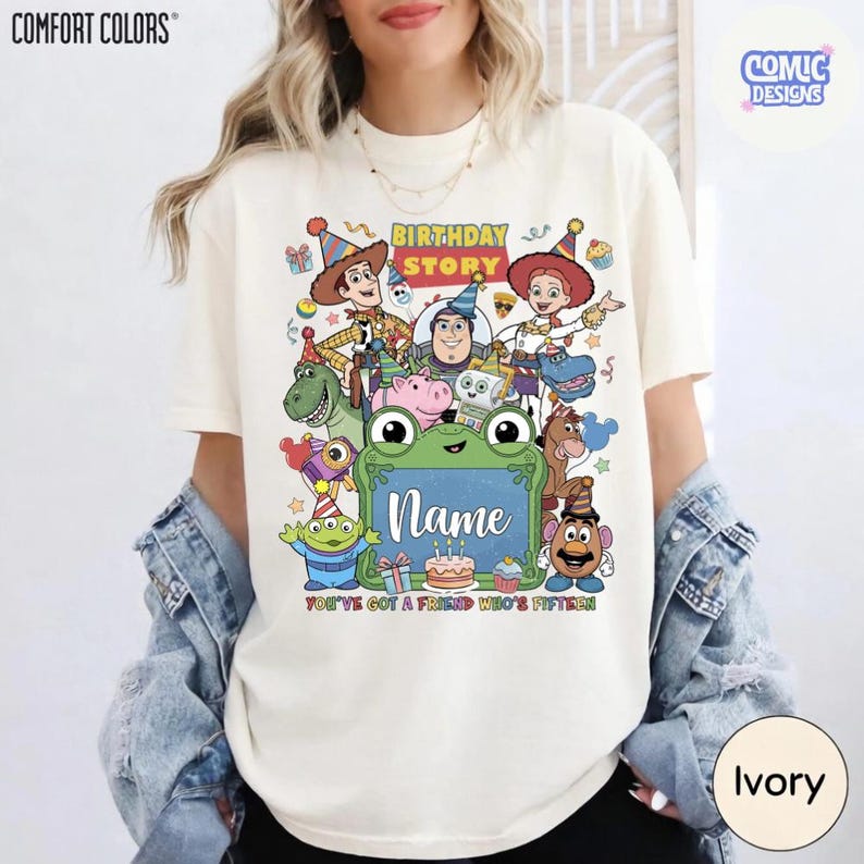 May include: An ivory t-shirt featuring a colorful graphic print of various animated characters wearing party hats. The design includes the text "BIRTHDAY STORY", "Name", and "YOU'VE GOT A FRIEND WHO'S FIFTEEN" along with illustrations of a cake, gifts, and a frog.