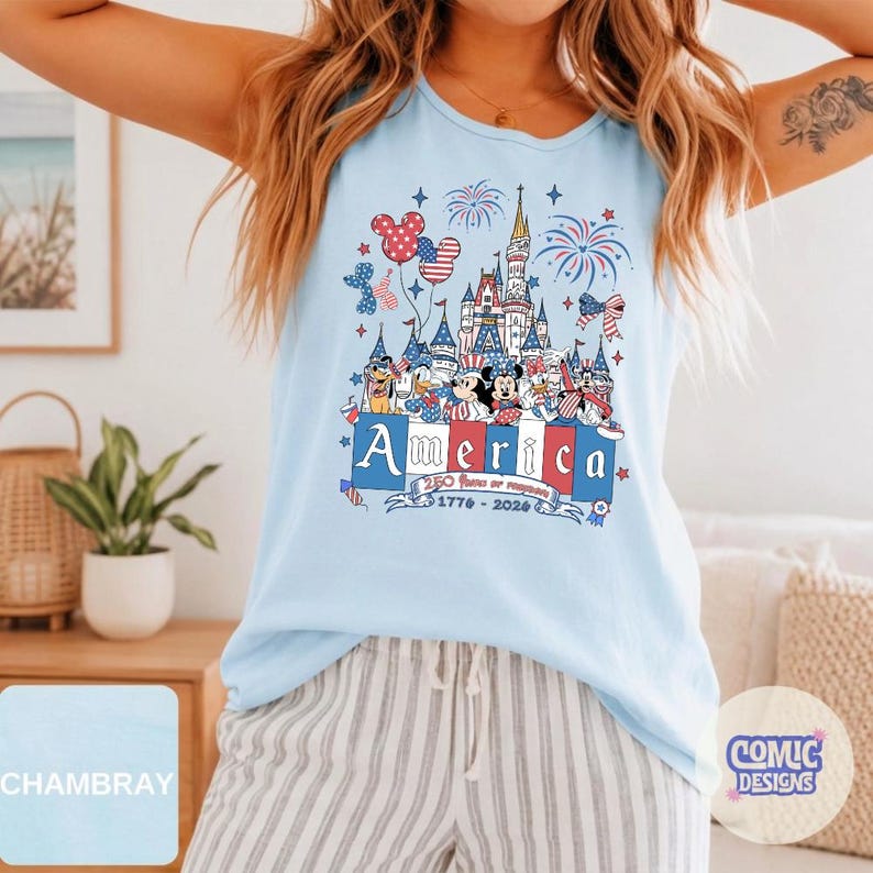 May include: A light blue sleeveless tank top featuring a graphic print of a castle, fireworks, and cartoon characters wearing patriotic red, white, and blue clothing. The text reads America, 250 Years of Freedom, and 1776-2026 in a banner.