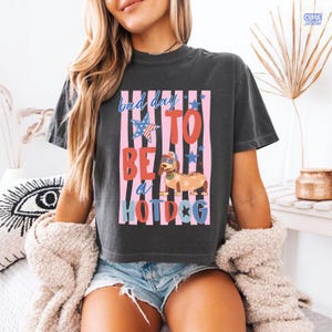 May include: Dark gray t-shirt with the phrase "Bad Day To Be a Hotdog" in a colorful design. The graphic includes a hotdog wearing sunglasses and a patriotic star. The shirt is a casual, comfortable style.