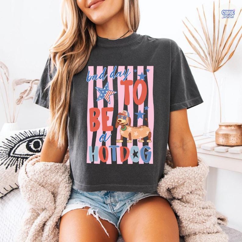 May include: Dark gray t-shirt with the phrase "Bad Day To Be a Hotdog" in a colorful design. The graphic includes a hotdog wearing sunglasses and a patriotic star. The shirt is a casual, comfortable style.