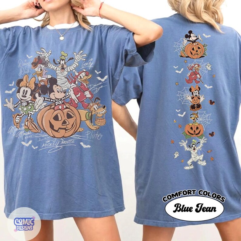 Op de afbeelding: Een blauw spijkerkleurig t-shirt met een grote grafische print van Disney-figuren in Halloween-kostuums op de voorkant en een verticale kolom met kleinere karakterillustraties op de achterkant, waaronder Mickey Mouse, Minnie Mouse, Donald Duck, Goofy en Pluto.