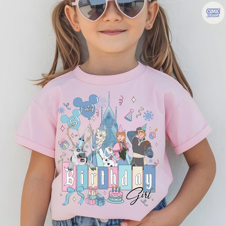 May include: Pink short-sleeved t-shirt featuring a colorful graphic of characters from a popular animated movie, including a reindeer, a snowman, and two princesses. The shirt has the text "Birthday Girl" and a number 5 balloon.