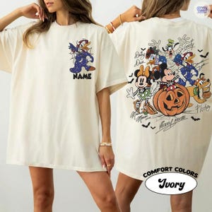 May include: An ivory t-shirt featuring a small graphic of Donald Duck in a costume on the front chest with the word NAME below it. The back displays a large Halloween-themed illustration of Mickey Mouse, Minnie Mouse, Goofy, Donald Duck, and Pluto.
